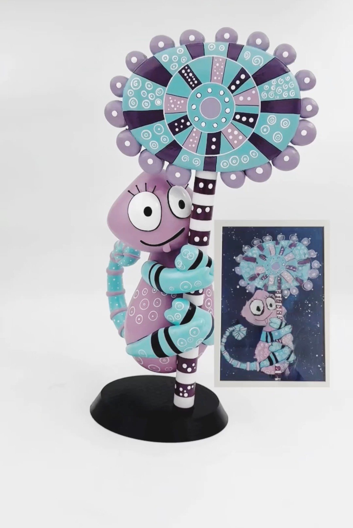 A purple and teal flower creature figurine beside the original painting