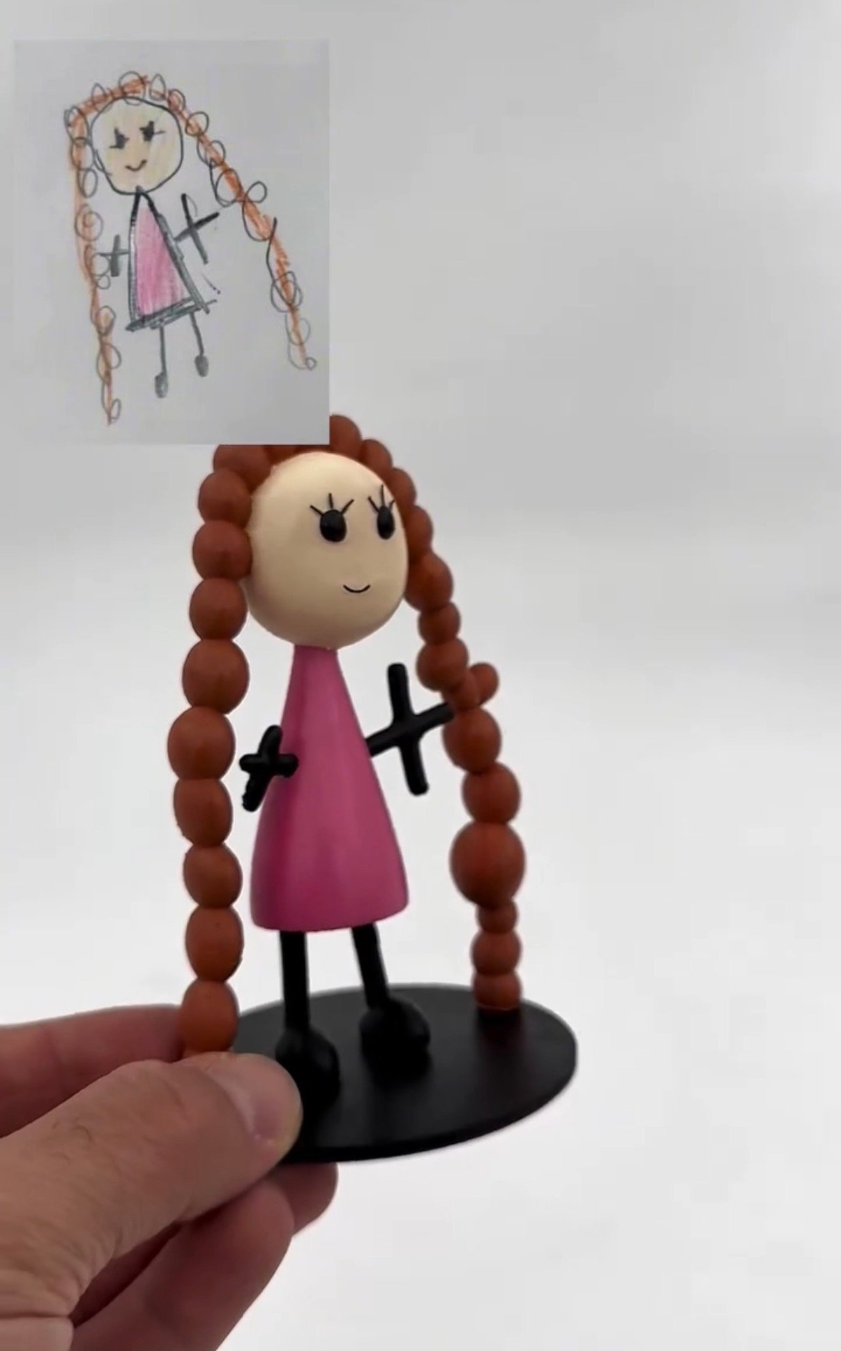 A figurine of a girl in a pink dress with beaded braids beside the original pencil drawing